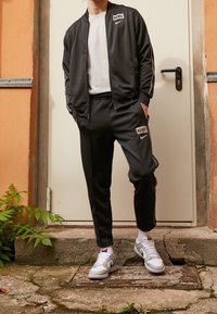 Tuta Nike nera con giacca con zip e pantaloni, caratterizzata da accenti e loghi bianchi. Indossata con sneakers bianche e grigie su una superficie di pietra.