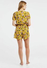 Combinaison fleurie en jaune moutarde, à manches courtes, avec une taille ceinturée, fabriquée dans un tissu doux présentant des fleurs roses et violettes ainsi que des feuilles vertes.