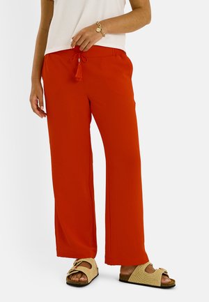 Personne portant un pantalon rouge ample avec une taille à cordon, un haut blanc, un bracelet doré et des sandales beige à enfiler avec des boucles.