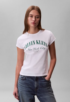 Młoda kobieta z prostymi jasnobrązowymi włosami nosi białą koszulkę z krótkim rękawem z zielonym napisem „Salvin Klein New York City” oraz niebieskie dżinsy, ręka w kieszeni.