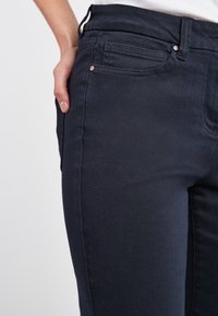 Detailní záběr lidské ruky odložené vedle přední kapsy tmavě námořnických kalhot na jednobarevném světlém pozadí.
