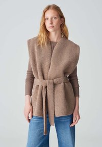 Beige Fleece-Weste mit Wickeldesign, ausgestattet mit einem breiten Kragen und einem Gürtel. Getragen über einem braunen Langarmoberteil und kombiniert mit blauen Jeans.