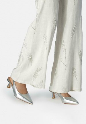 Tacones slingback plateados de punta estrecha, usados con pantalones blancos de pierna ancha que tienen sutiles patrones bordados de hojas.