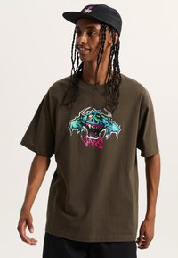 Camiseta de algodón marrón con mangas cortas, que presenta un gráfico de un monstruo colorido en azul y rosa, combinada con una gorra negra.