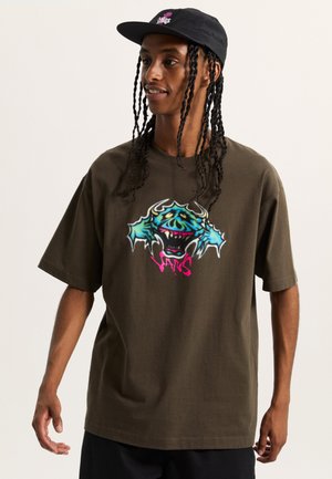 Brown cotton t-shirt with short sleeves, featuring a colorful monster graphic in blue and pink, paired with a black cap.