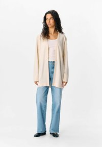 Cardigan beige chiaro a costine, con maniche lunghe, apertura frontale e vestibilità comoda, abbinato a jeans a palazzo blu chiaro e scarpe nere.