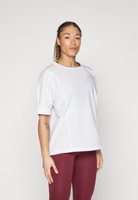Valge, lühikeste varrukatega sporttop ümara kaelusega, millel on sile ja pehme tekstuur ning diskreetne logoelement. Kombineeritud burgundi värvi retuusidega.
