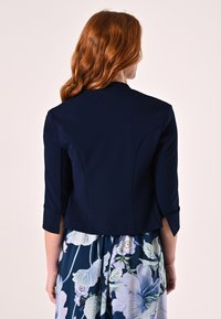 Veste cropped navy à la coupe cintrée, manches trois-quarts et texture lisse ; associée à une robe à motifs floraux.