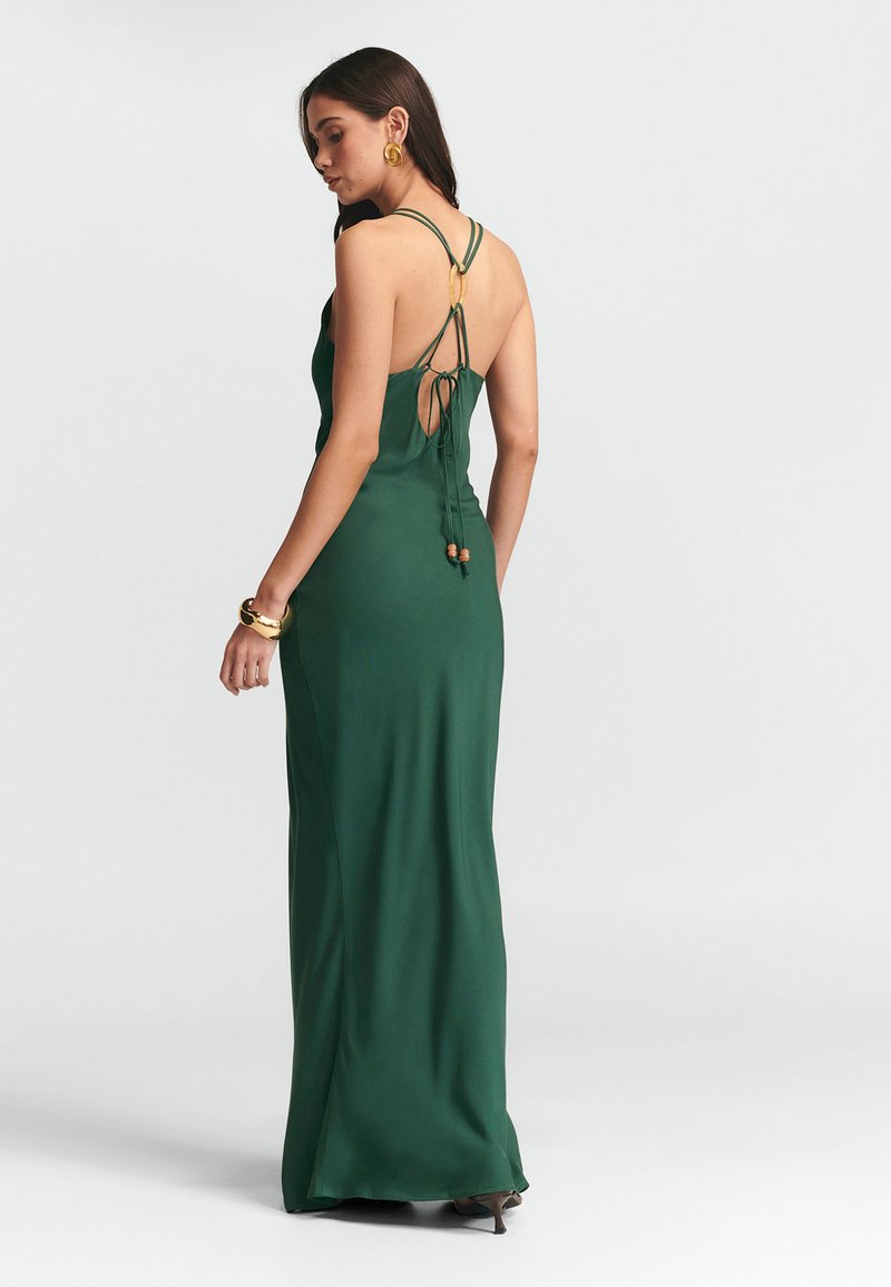 Lång grön satinklänning med djup rygg, tunna korsettselar och dekorativa band. Slät textur och en flödande silhuett.