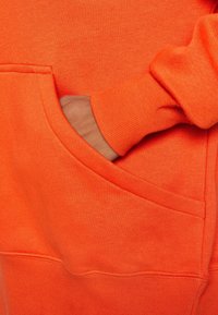 Orange sweatshirt med en framficka. Har ribbad mudd och fåll, en mjuk textur och en enkel, avslappnad design.