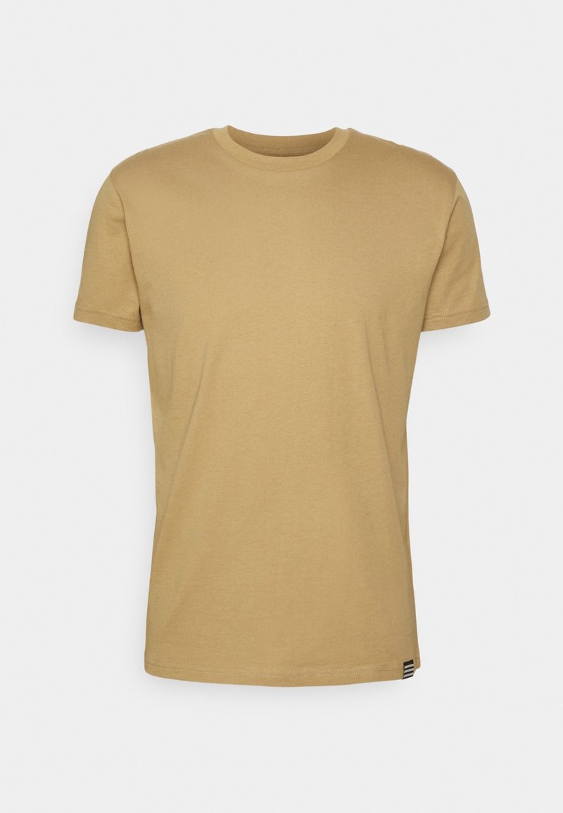T-shirt beige à manches courtes et col rond avec une petite étiquette rayée sur l'ourlet du bas, sur un fond uni gris clair.