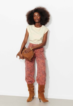 Femme avec une grande afro portant un haut beige sans manches, un pantalon rose à fleurs, des bottes à talons en daim marron, tenant un sac assorti en daim marron.