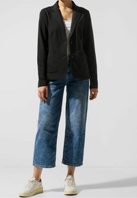 Černý blazer z bavlněné směsi s klopovým límcem a dvěma předními kapsami, doplněný o modré džínsy s vysokým pasem a širokými nohavicemi a smetanové tenisky.