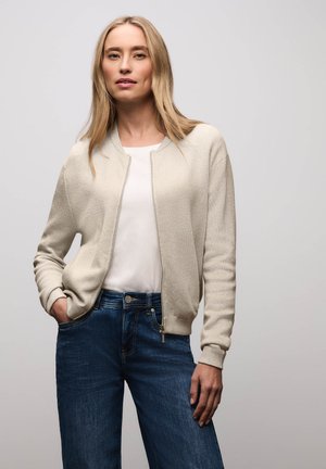 Blondynka w jasnobeżowym rozpinanym kardiganie na zamek, pod którym ma białą koszulę i niebieskie dżinsy, stoi z jedną ręką w kieszeni.