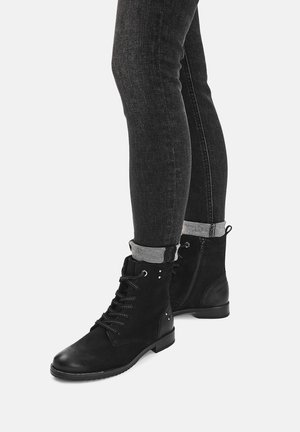 Fekete felgöngyölített skinny farmert viselő lábak fekete cipőfűzős bokacsizmában, oldalzippel, világos háttér előtt.