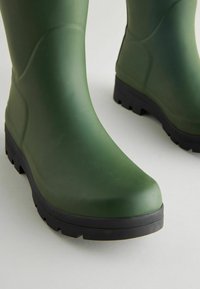 Groene rubberen regenlaarzen met een gladde afwerking, een ronde neus en een zwart patroon op de zool. De bovenkant heeft een licht verhoogd ontwerp.