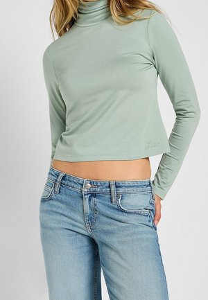 Nainen, joka pukeutunut vaaleanvihreään pitkähihainen poolokauluksiseen lyhytpaita ja vaaleansinisiin korkeavyötäröisiin farkkuihin, joissa on metallinen nappisuljenta.