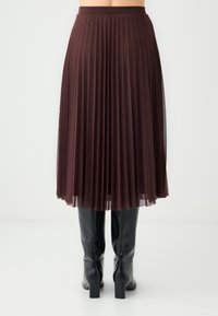 Jupe plissée marron avec un voile transparent, longueur mi-mollet, taille élastique, portée avec des bottes noires montantes jusqu'aux genoux.