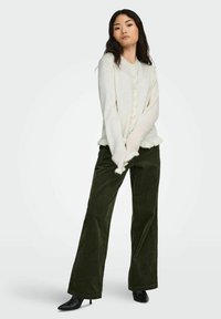 Cardigan beige chiaro con dettagli di bottoni e orlo arricciato abbinato a pantaloni a gamba larga in corduroy verde scuro; modella che indossa stivali neri alla caviglia.