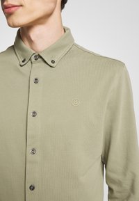 Olivově zelená košile na knoflíky z bavlněné látky s límečkem, se černými knoflíky a malým béžovým logem na hrudi.