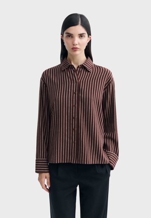 Femme aux cheveux raides et foncés portant une chemise marron à boutons avec des rayures verticales blanches et un pantalon noir, debout devant un fond uni.