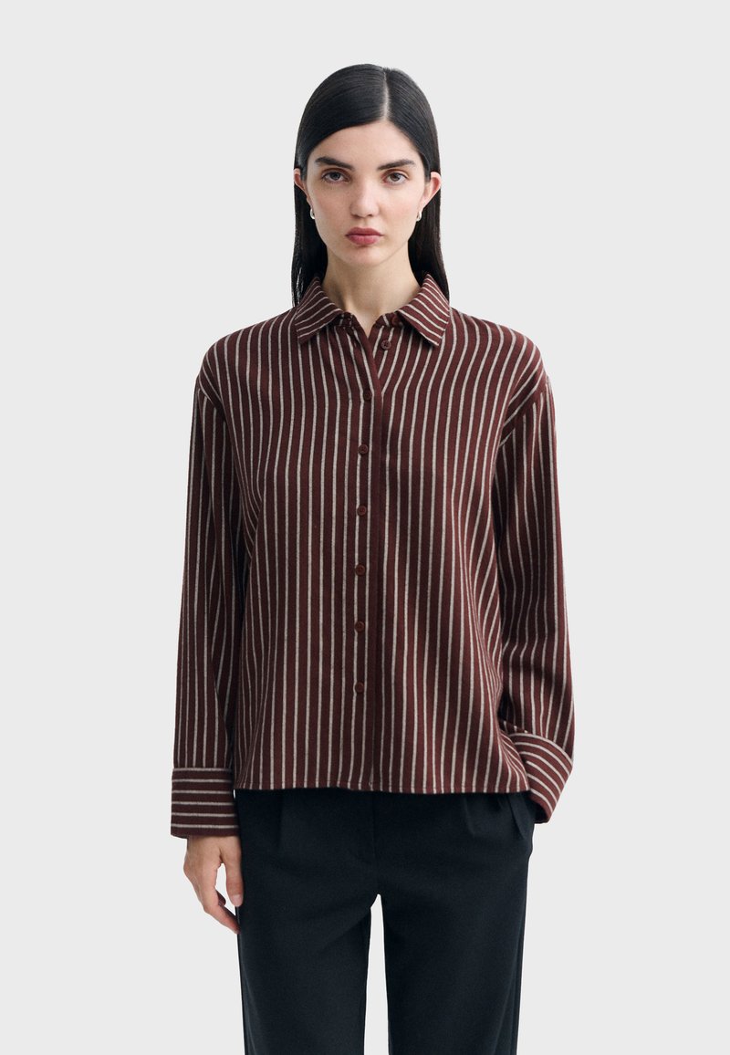 Femme aux cheveux raides et foncés portant une chemise marron à boutons avec des rayures verticales blanches et un pantalon noir, debout devant un fond uni.