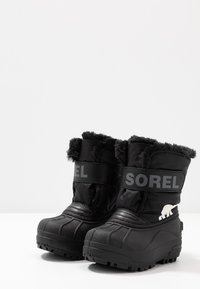 Botas de invierno negras con forro de piel sintética, suelas de goma y marca Sorel. Presentan un acento de oso polar blanco en el talón.
