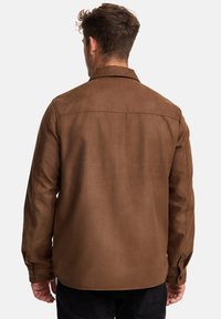 Homme de dos portant une chemise-veste marron à manches longues et un pantalon noir, devant un fond clair.