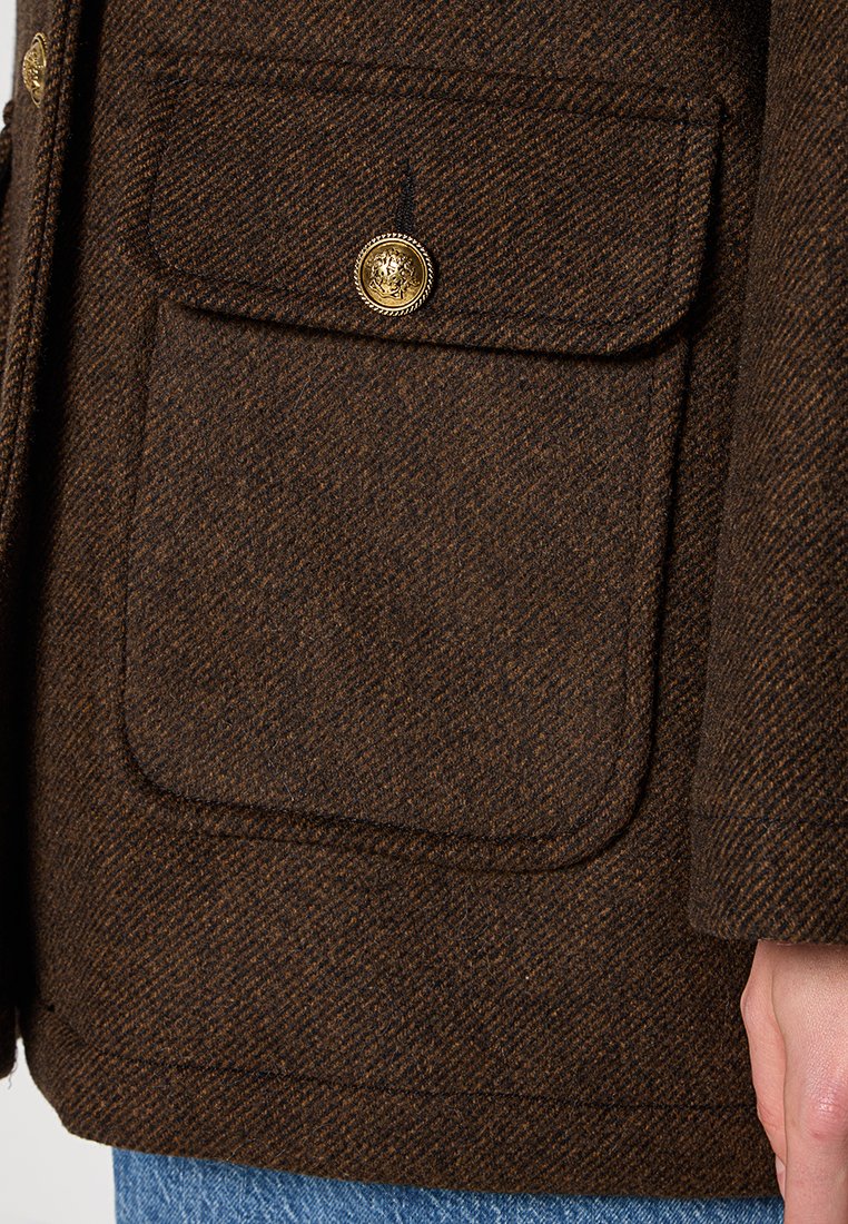 Close-up van een bruine wollen jaszak met een gouden knop, gedragen met blauwe jeans en de hand van de drager aan de zijkant.