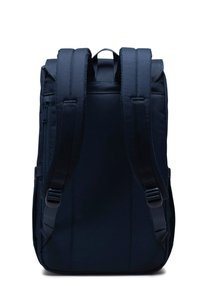 Marinblå ryggsäck med rektangulär form, vadderade axelremmar och ett handtag på toppen. Materialet verkar hållbart och texturerat, med sidofickor med dragkedja.