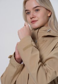 Beige wasserdichte Jacke mit einem Frontreißverschluss, verstellbarem Kordelzug-Kapuze und Klettverschlüssen an den Ärmelbündchen. Glatte Textur und minimalistisches Design.