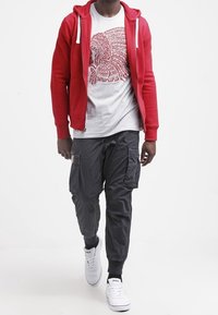 Röd zip-up hoodie över en grå grafisk t-shirt. Svarta cargobyxor med flera fickor. Vita sneakers med svarta detaljer.