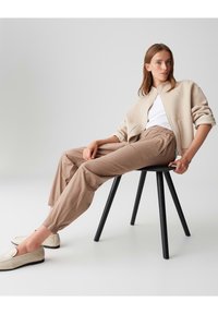 Beige Wollmischung-Jacke über einem weißen T-Shirt, kombiniert mit hellbraunen schmalen Hosen und cremefarbenen Slippern, sitzt auf einem schwarzen Holzstuhl.