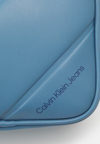 Borsa in faux leather blu con un design diagonale. Logo "Calvin Klein Jeans" impresso in blu scuro. Texture liscia e forma strutturata.