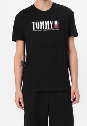 Henkilö, joka käyttää mustaa Tommy Jeans -t-paitaa, jossa on valkoinen, punainen ja sininen logo sekä teksti "TOMMY JEANS WORLDWIDE SINCE 1985" rinnassa.