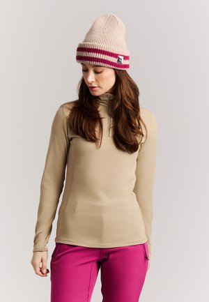 Kvinde med langt brunt hår iført en beige langærmet top, beige strikket hue med røde striber og lyserøde bukser mod en ensfarvet baggrund.