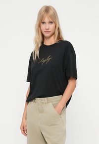 Blond naine musta T-särgiga, millel on kuldne kiri, ja beežide pükstega, seismine ühe käega taskus valge tausta ees.