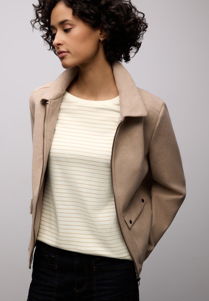 Beige Reißverschlussjacke mit strukturierter Oberfläche, mit Stehkragen und Seitentaschen, getragen über einem cremefarbenen Oberteil mit dünnen gelben Streifen.