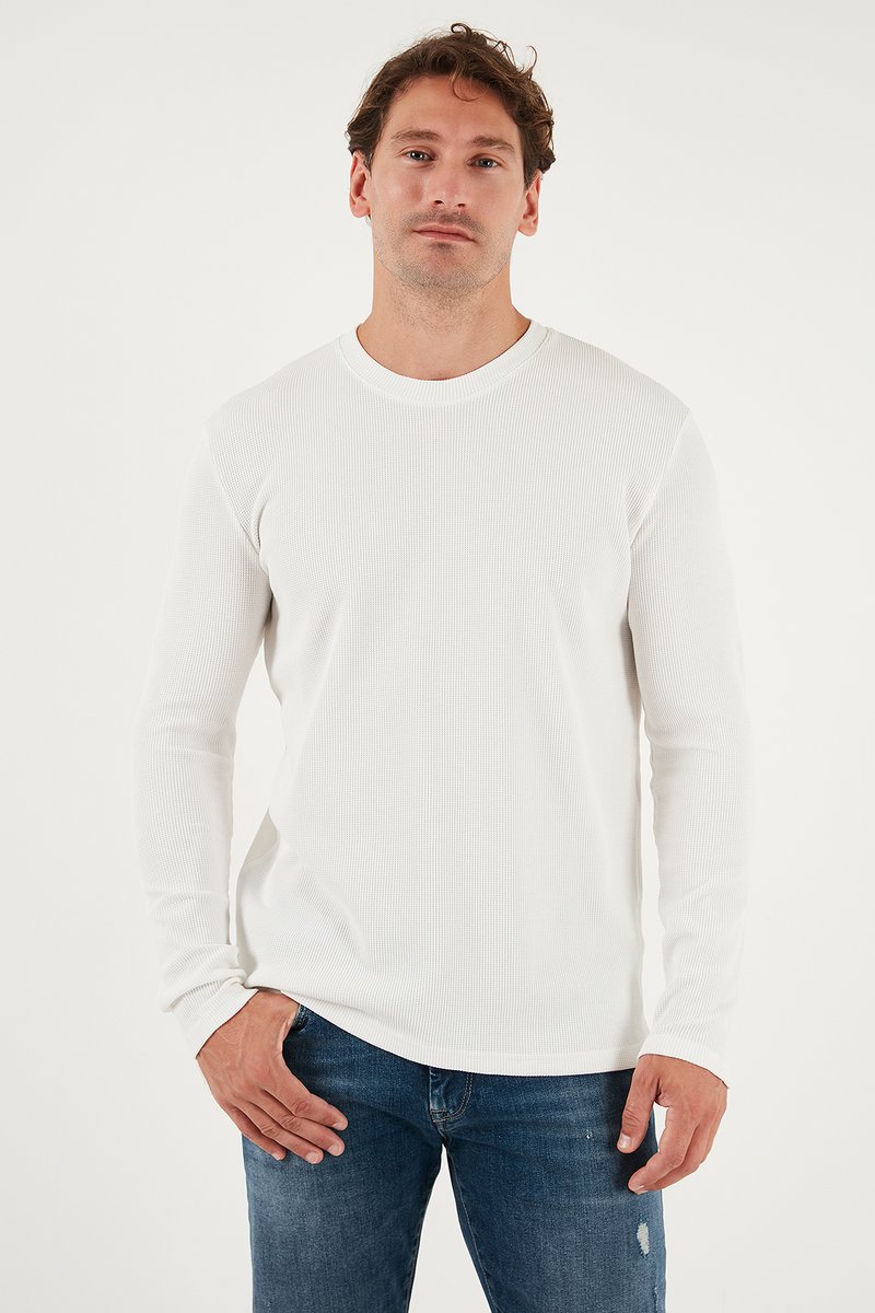 T-shirt blanc à manches longues en côte avec un col rond, porté avec un jean bleu déchiré, présentant un tissu texturé et une coupe décontractée.