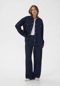 Mörk denimjacka med två bröstfickor, knäppningar och avslappnad passform, kombinerad med matchande vida byxor och en vit t-shirt.