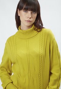 Strickpullover in Gelb mit hohem Rippkragen, ausgestattet mit strukturierten Zopfmustern und einer lockeren Passform. Der Stoff wirkt weich und warm.