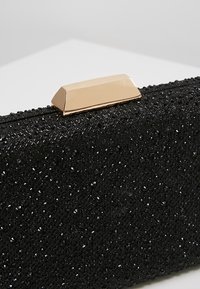 Gros plan d'une pochette noire texturée avec de petites perles noires et un fermoir rectangulaire doré sur une surface blanche.