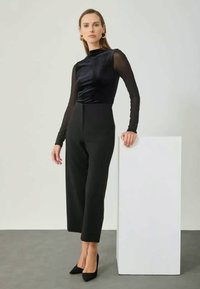 Black long-sleeve top with a smooth finish paired with high-waisted black trousers, set against a neutral background, accentuated by pointed heels.