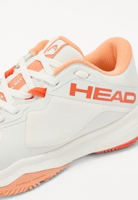 Bijela sportska cipela s teksturiranim gornjim dijelom, narančastim detaljima i istaknutim logotipom "HEAD" sa strane. Ima kopčanje na vezanje i gumeni potplat.