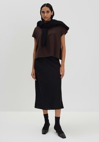 Braunes, kurzärmeliges Oberteil über einem schwarzen Midirock, mit einem gestrickten schwarzen Schal, der um die Schultern drapiert ist. Schwarze, strukturierte Socken und Schuhe mit quadratischer Zehenpartie.