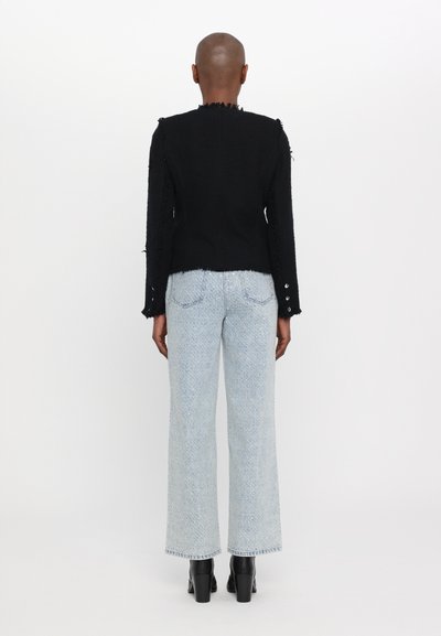 Schwarze Textiljacke mit ausgefransten Kanten, kombiniert mit hellblauen weitem Jeans. Das Outfit weist Knopfakzente an den Ärmeln auf.