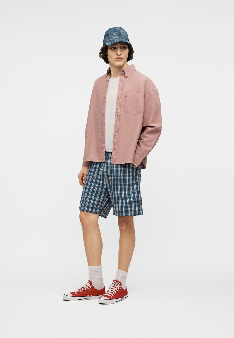 Junger Mann, der eine blaue Jeansmütze, ein rosa Hemd mit Knöpfen, ein weißes T-Shirt, blaue karierte Shorts, weiße Socken und rote Turnschuhe trägt, vor einem einfarbigen Hintergrund steht.