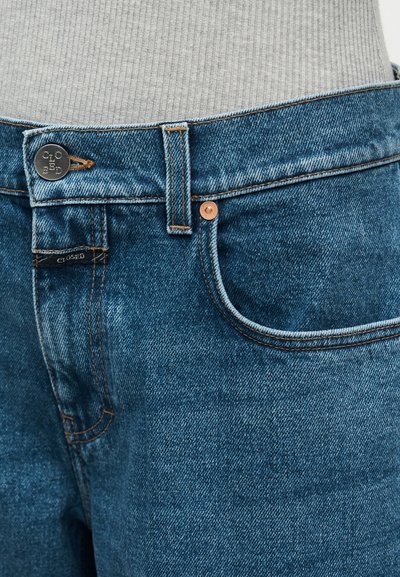 Μπλε τζιν παντελόνια με μεσαία μέση, χαλκοκόκκινα κουμπιά και αντίθετη ραφή. Διαθέτουν μπροστινή τσέπη και υφή φινιρίσματος. Ορατή ετικέτα μάρκας.