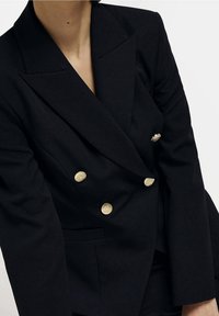 Kvinna iklädd en svart dubbelknäppt blazer med guldknappar, med breda slag och strukturerade axlar.