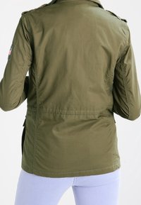 Kurtka oliwkowa wykonana z miękkiego, trwałego materiału. Charakteryzuje się dopasowanym krojem, pagonami na ramionach oraz taliowaniem.